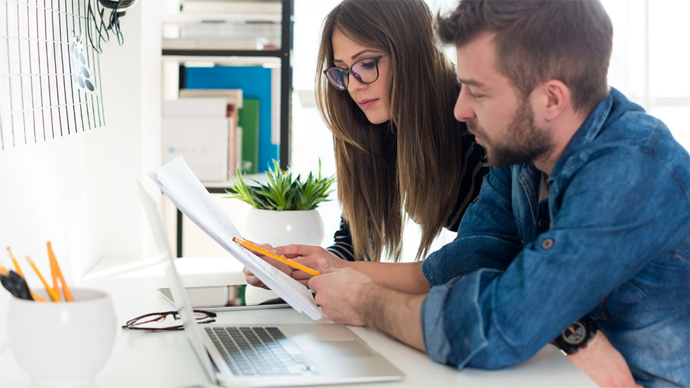 a man and woman looking over some papers while sitting in front of a laptop